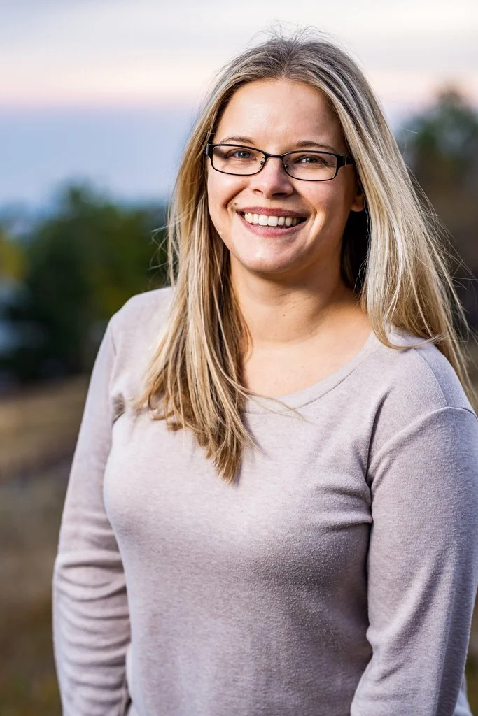 A young woman with long blonde hair and glasses smiling outdoors.