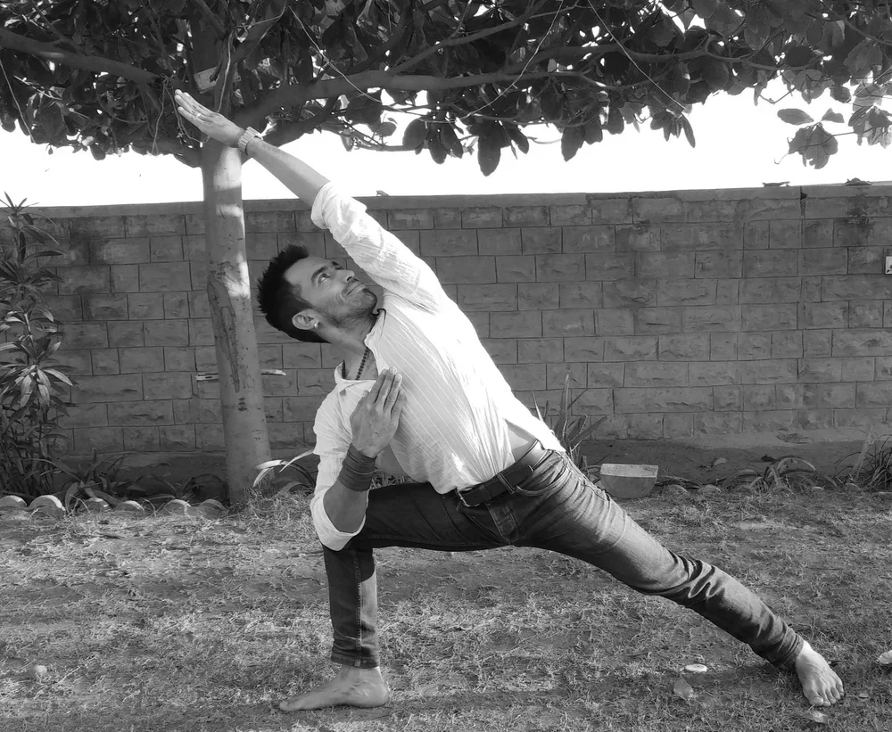 Un homme en position de yoga dans un jardin, effectuant une posture de guerrier, en noir et blanc.