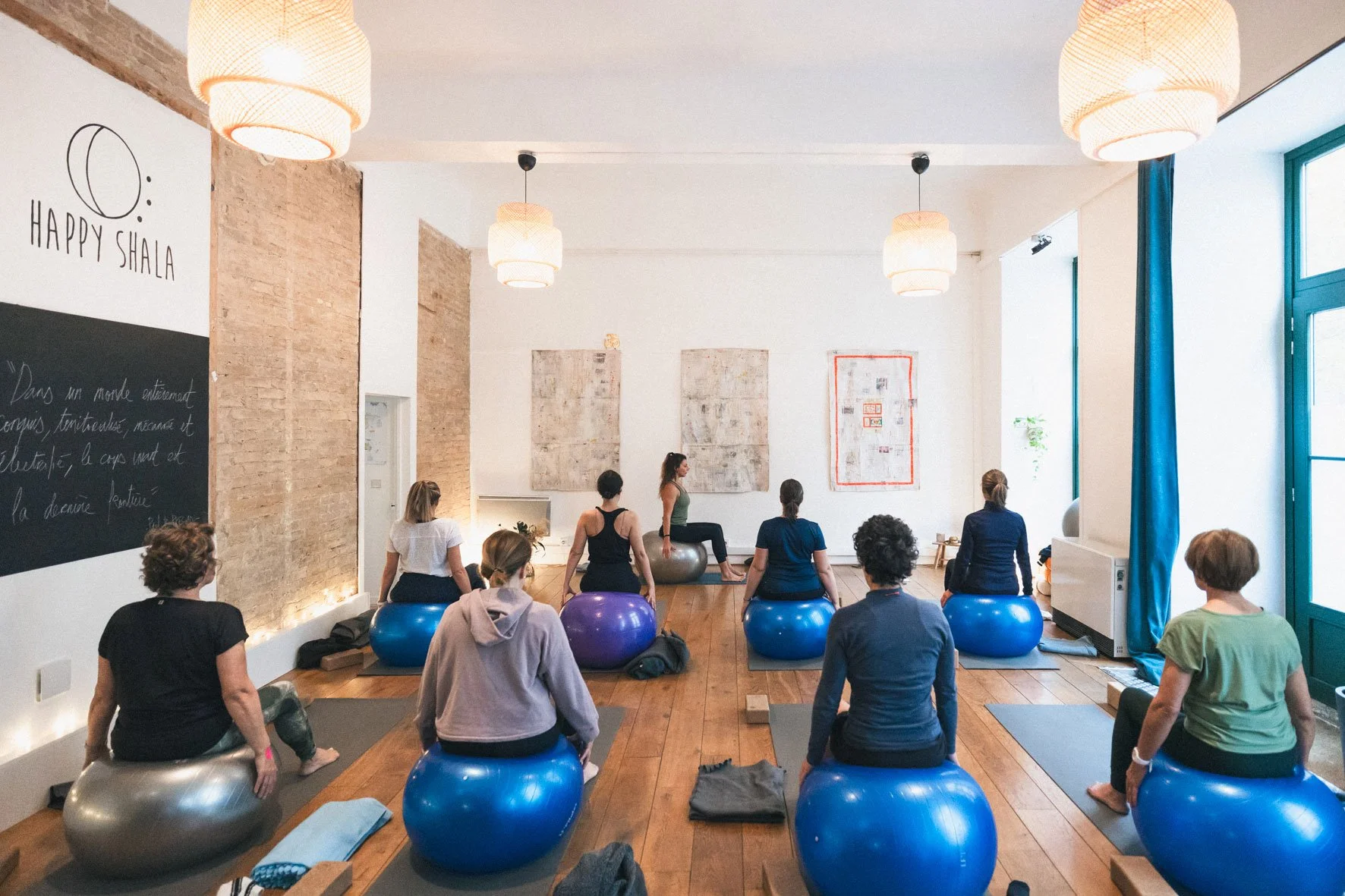 Cours de yoga ou méditation dans une salle lumineuse avec des participants assis sur des ballons de fitness, face à un instructeur en position de méditation ou de yoga. La salle a des murs blancs, des œuvres d'art suspendues, des lampes suspendues, et une grande fenêtre avec des rideaux bleus.