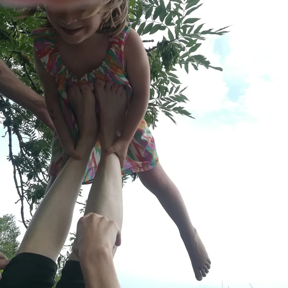 Une fille en robe colorée est soulevée dans les airs par des mains, avec un ciel clair et des arbres en arrière-plan.