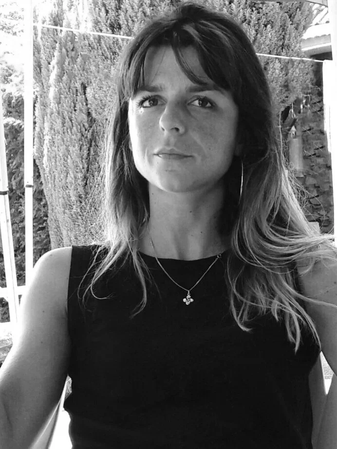 Une femme avec des cheveux mi-longs, portant un débardeur noir et un pendentif, prise en photo en extérieur avec un arbre en arrière-plan.