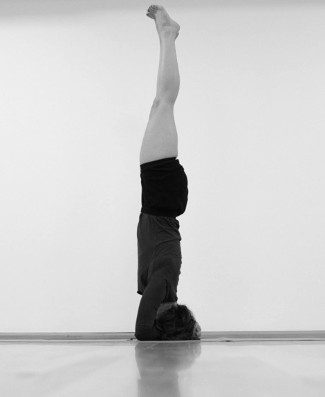 Personne pratiquant la yoga en position de tête, bras pliés, dans une pièce avec un sol en bois.