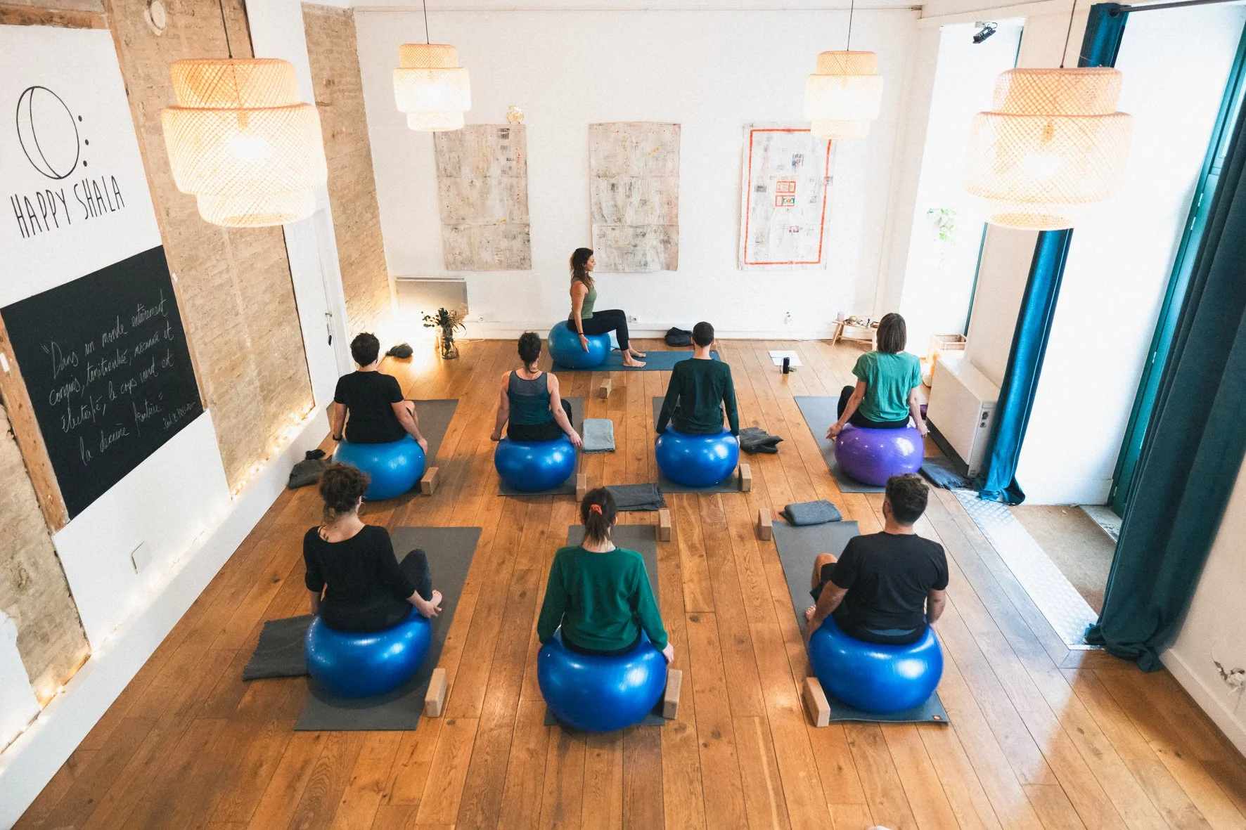 Cours de yoga dans un studio lumineux avec des participants assis sur des ballons d'exercice, une instructrice en position debout à l'avant, mur blanc avec des œuvres d'art, lampes suspendues en plafond, sol en bois. Happy Shala Studio Yoga Pilates G