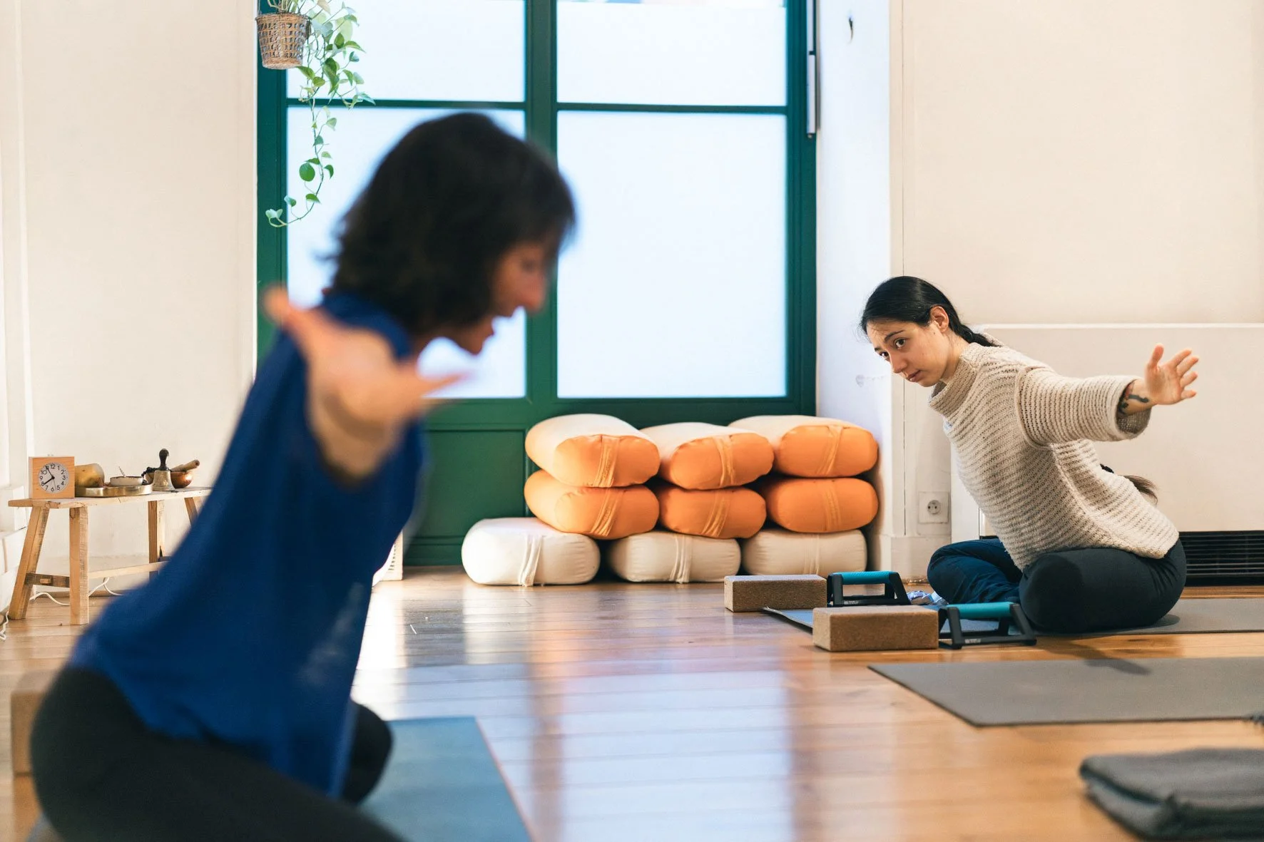 Deux femmes pratiquant le yoga ou la méditation dans une salle lumineuse avec des coussins orange empilés en arrière-plan. L'une d'elles est en position de yoga, l'autre en pleine séance de méditation ou étirement. Happy Shala Studio Yoga Pilates Gre
