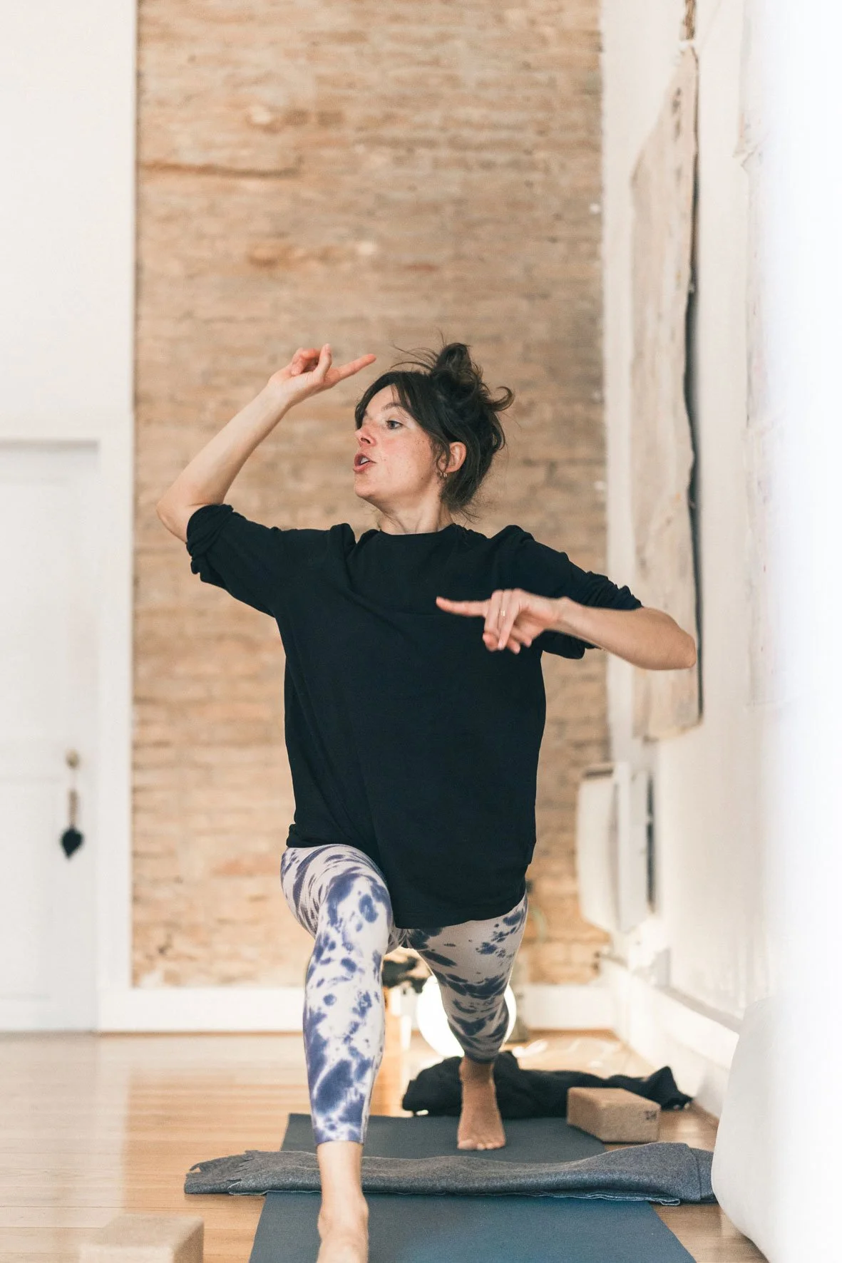 Une femme pratique le yoga ou la méditation en équilibre sur un pied dans une pièce avec un mur en briques, portant un t-shirt noir et des leggings à motif. Happy Shala Studio Yoga Pilates Grenoble