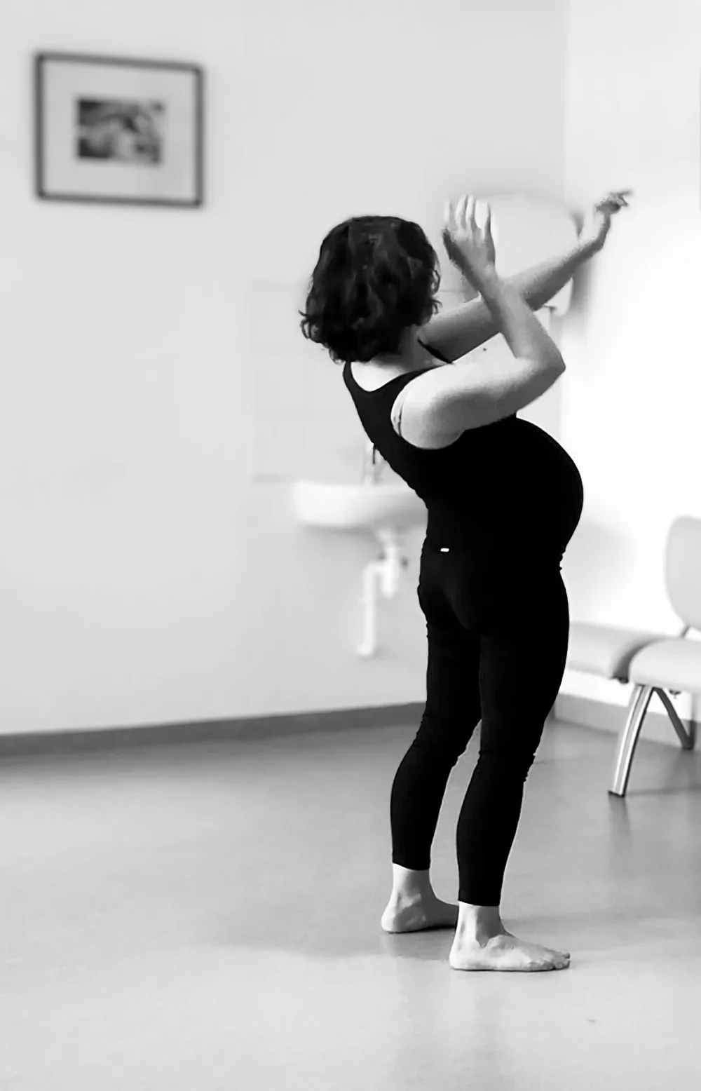 Une femme enceinte en train de faire de la danse ou du yoga dans une pièce blanche, avec une chaise et un tableau en arrière-plan, en noir et blanc.