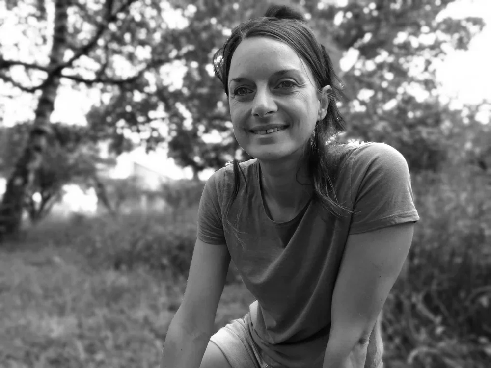 Une femme souriante assise dans un parc, entourée d'arbres et de nature, en noir et blanc.