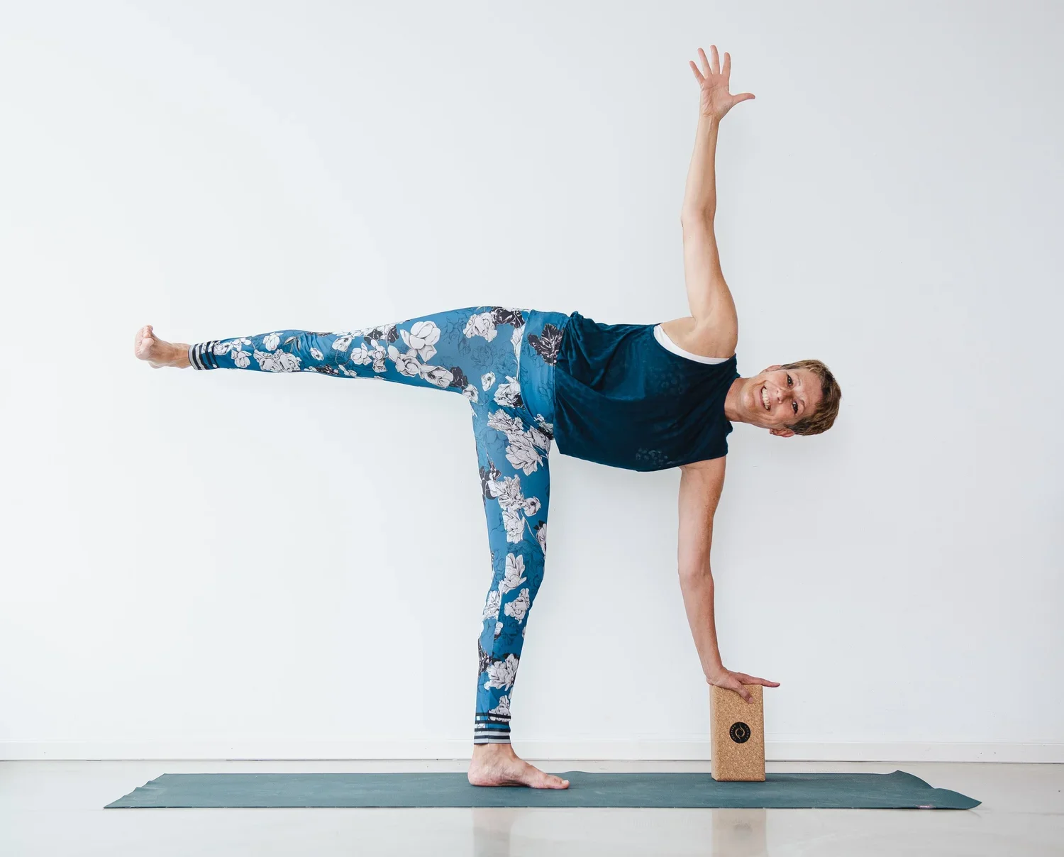 Homme faisant du yoga en équilibre avec un bloc en appui contre le sol, portant un haut noir et un legging bleu avec des motifs floraux.