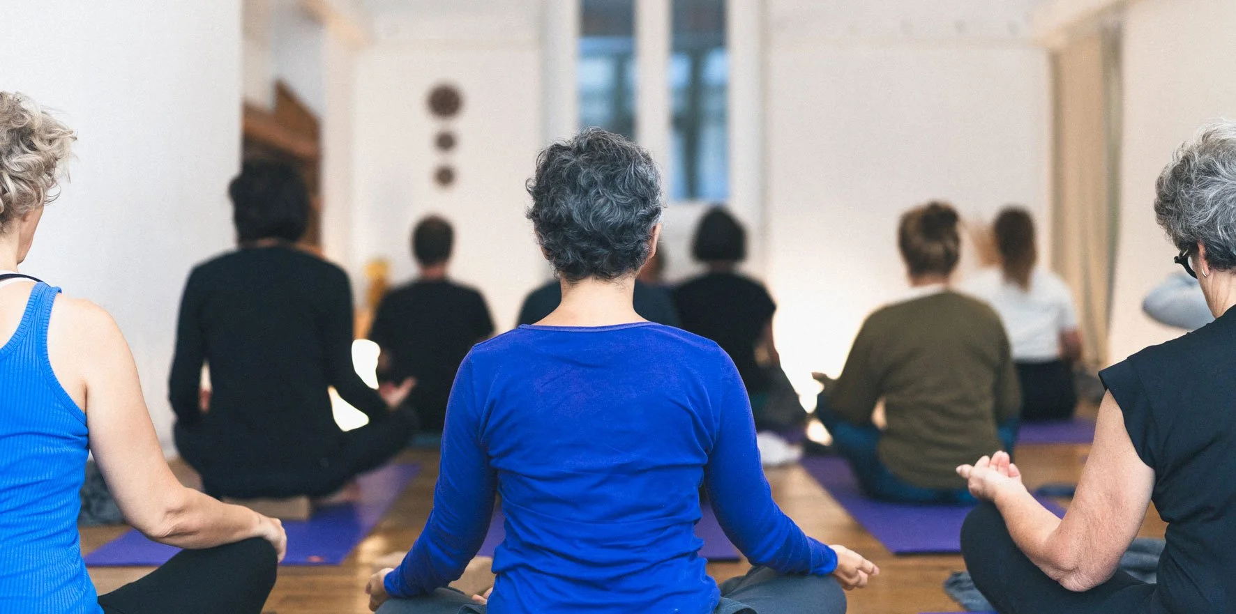 Groupe de personnes pratiquant la méditation ou le yoga en intérieur, assises sur des tapis, face à un instructeur devant un mur blanc. Happy Shala Studio Yoga Pilates Grenoble