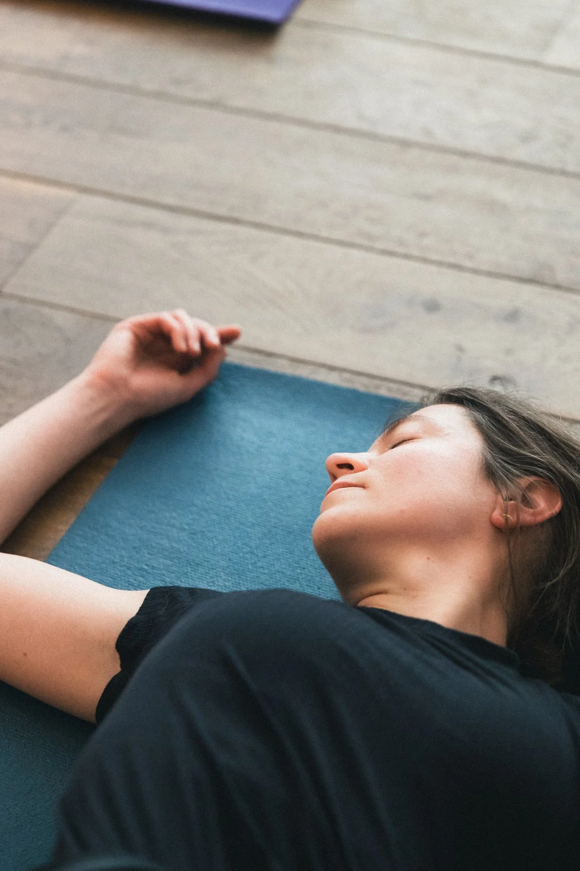 Une femme endormie allongée sur un tapis de yoga bleu, posée sur un sol en bois, dans une pièce lumineuse.