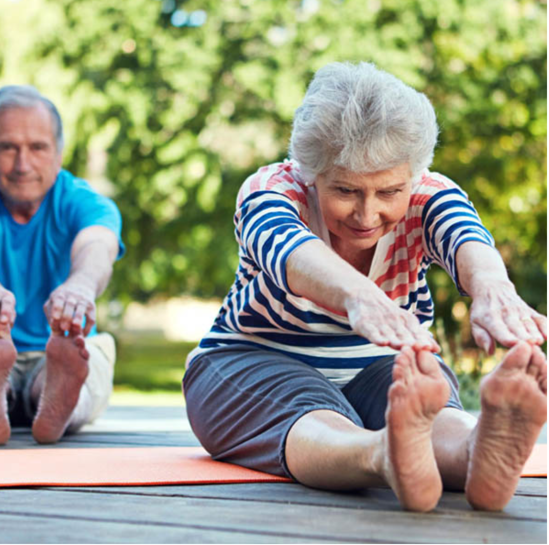 Deux personnes âgées faisant des étirements ou du yoga sur une terrasse en plein air, entourées d'arbres verts.