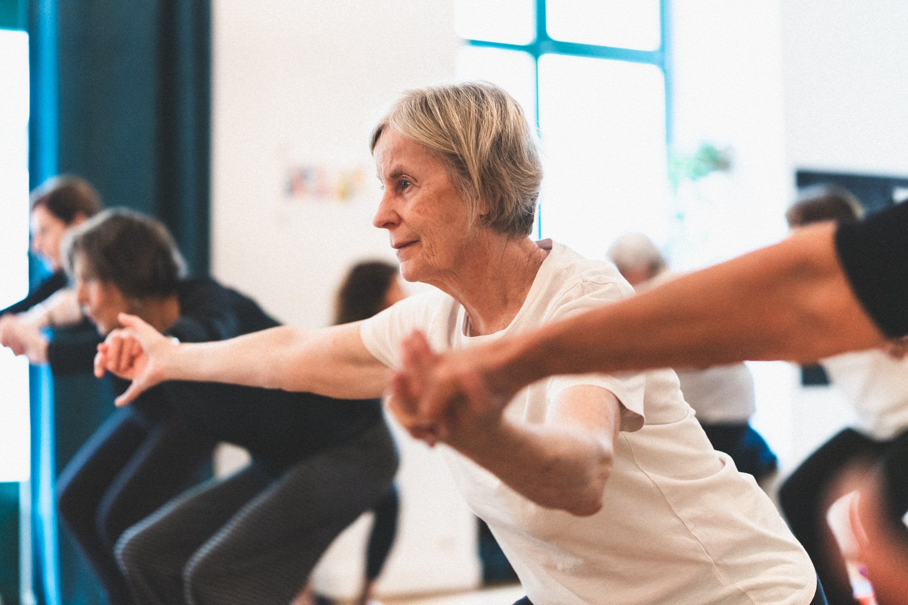 Une femme âgée participe à une séance de yoga ou d'exercice de groupe dans une salle lumineuse, étirant son bras en avant avec d'autres personnes derrière elle. Happy Shala Studio Yoga Pilates Grenoble