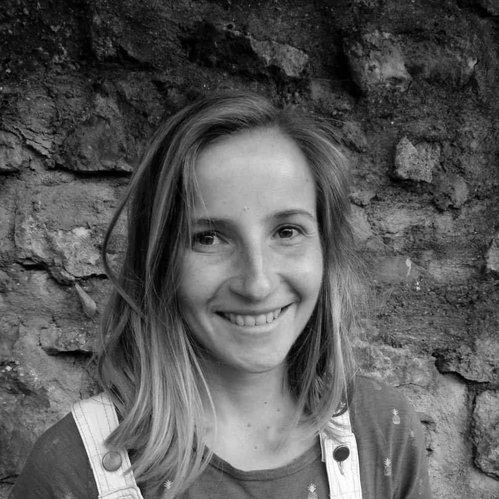 Portrait d'une jeune femme souriante avec des cheveux longs et ondulés, en maillot à bretelles, devant un mur en pierre. Happy Shala Studio Yoga Pilates Grenoble