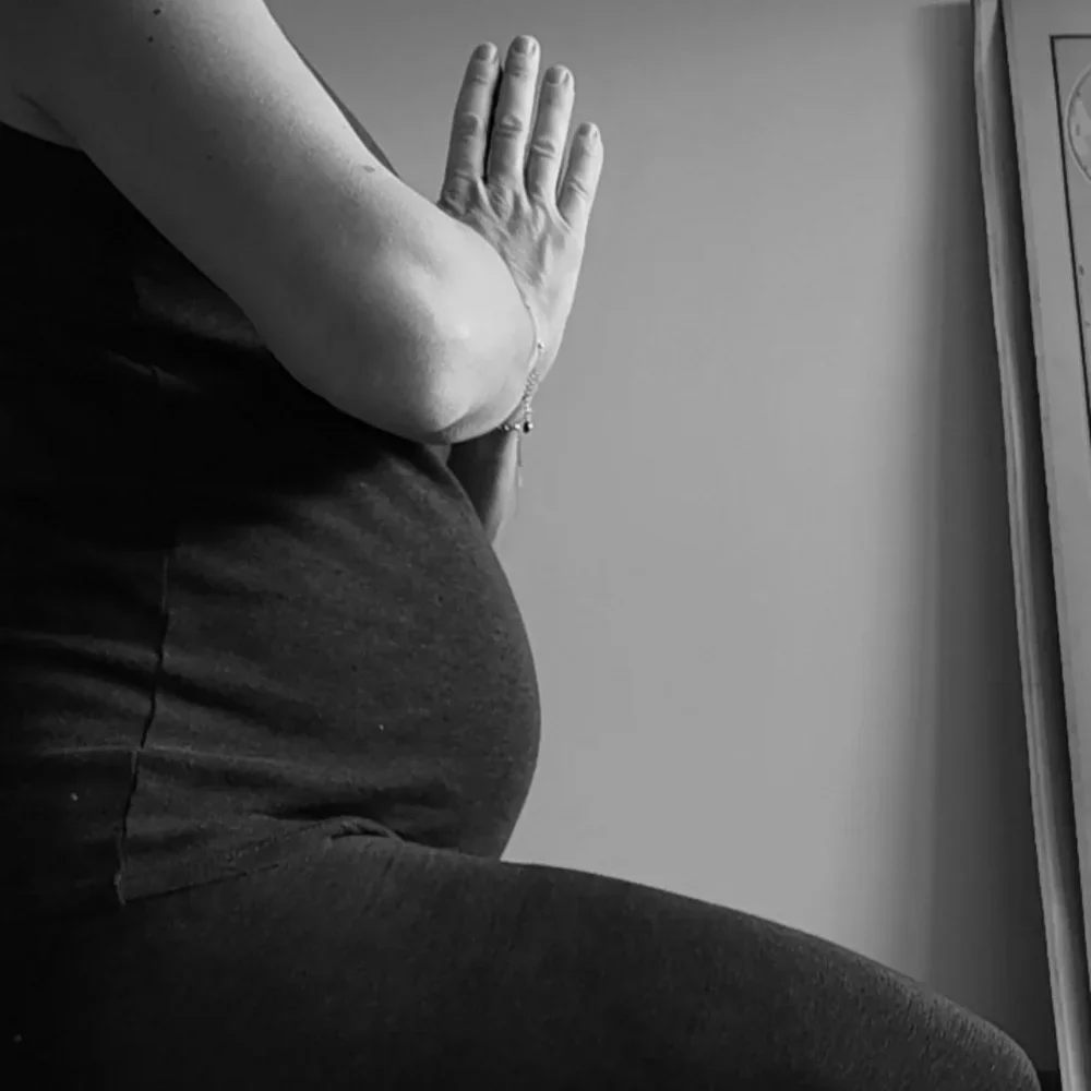 Photo en noir et blanc d'une personne enceinte assise avec une main posée sur son ventre, dans un environnement intérieur. Happy Shala Studio Yoga Pilates Grenoble