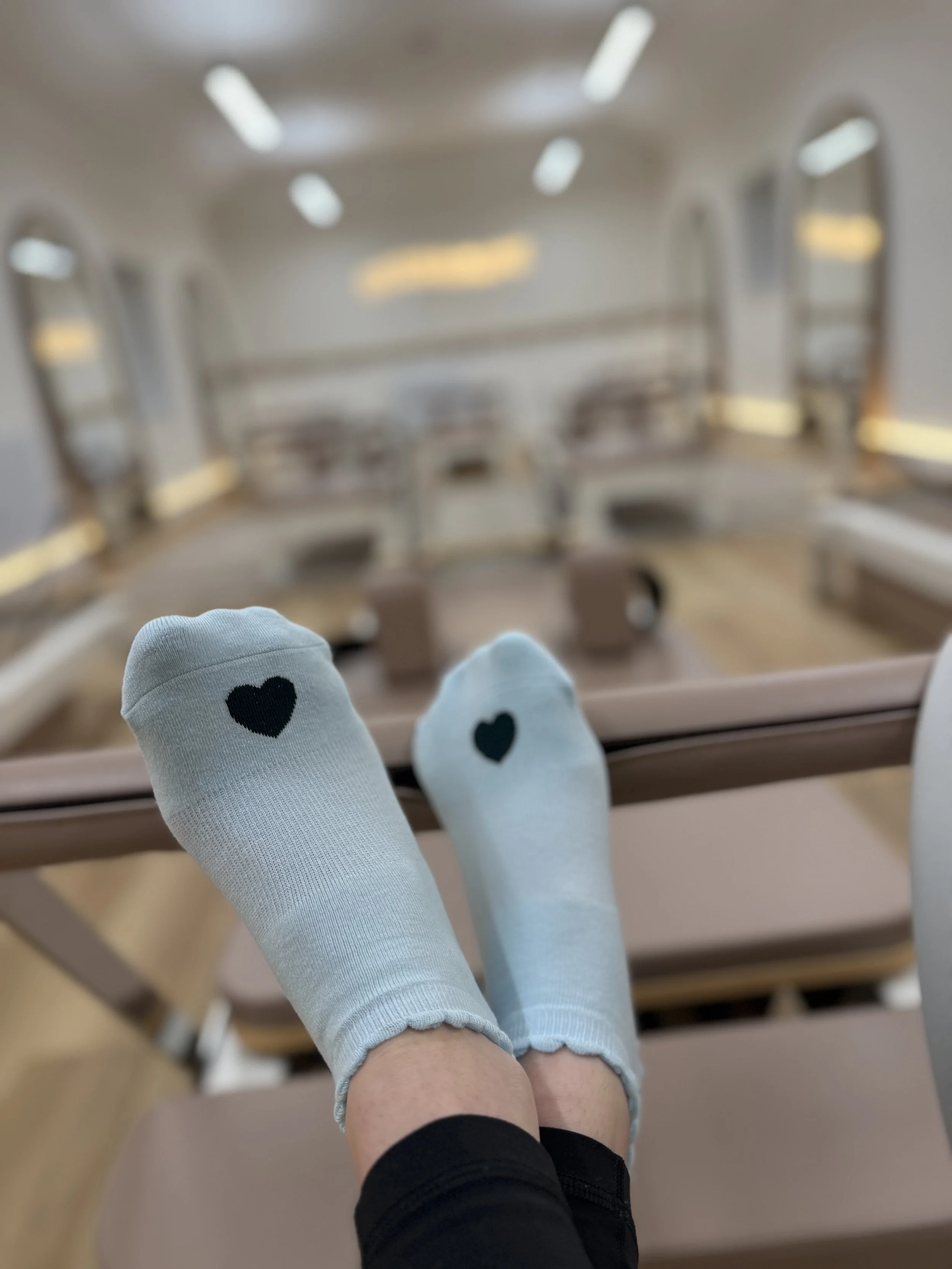 Person wearing white socks with black heart design, sitting on a wooden bench in a Pilates studio with white walls and recessed lighting.