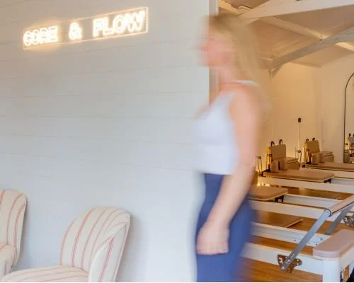 Blurred image of a woman standing in a room with a neon sign that reads 'CORE & FLOW' on the wall, with chairs in the background.
