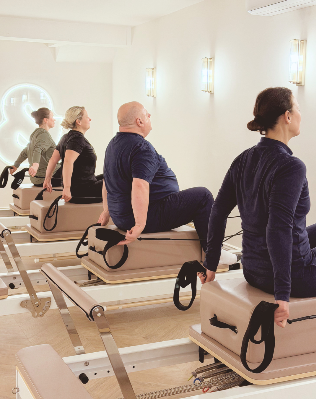 	
People participating in a Pilates or reformer exercise class in a studio, seated on Pilates reformer machines, holding handles with resistance bands at Core & Flow Thirs