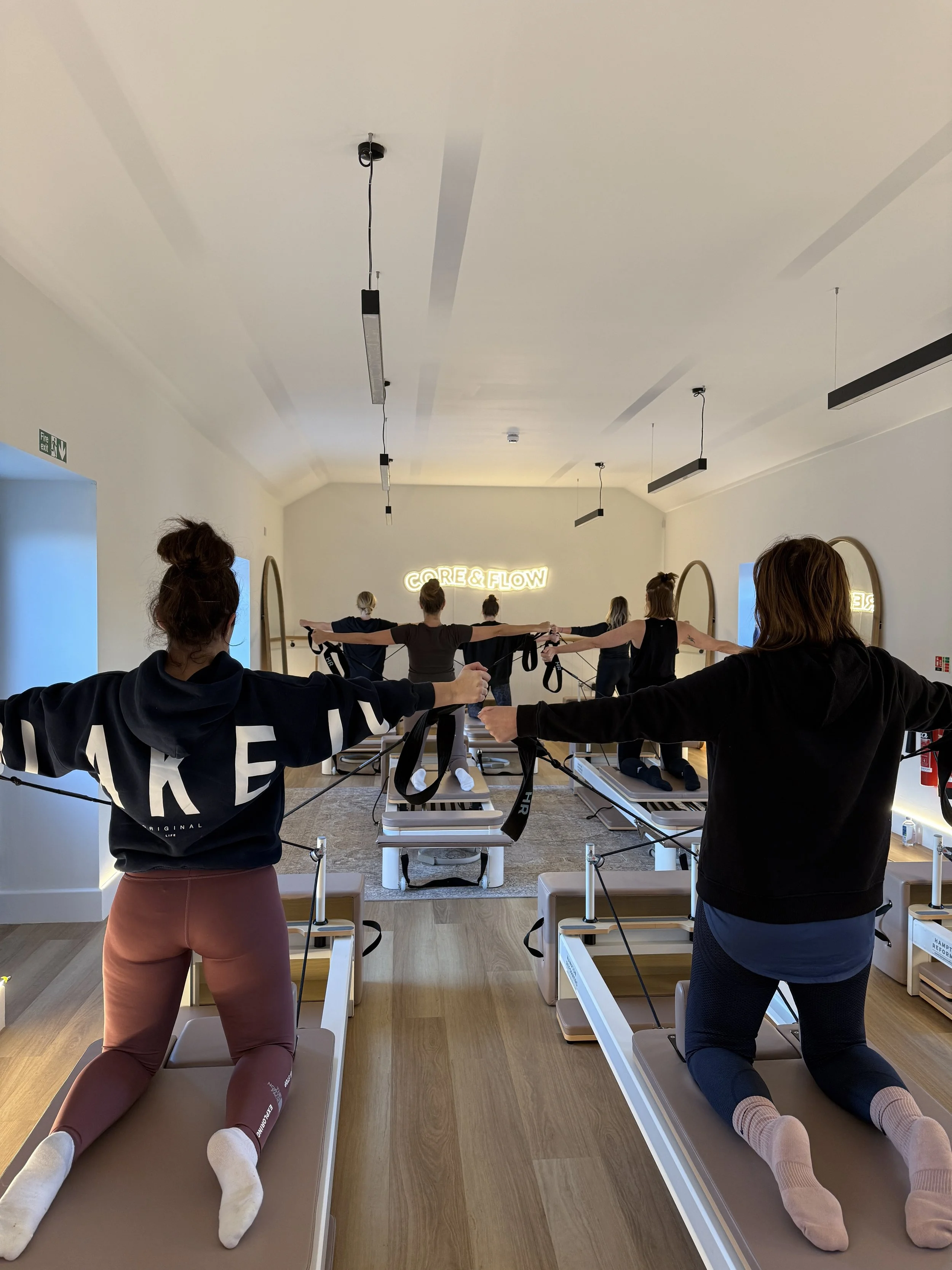 Group of people participating in a Pilates class, using Pilates reformer machines in a studio with a illuminated neon sign that reads 'CORE & FLOW' on the wall.