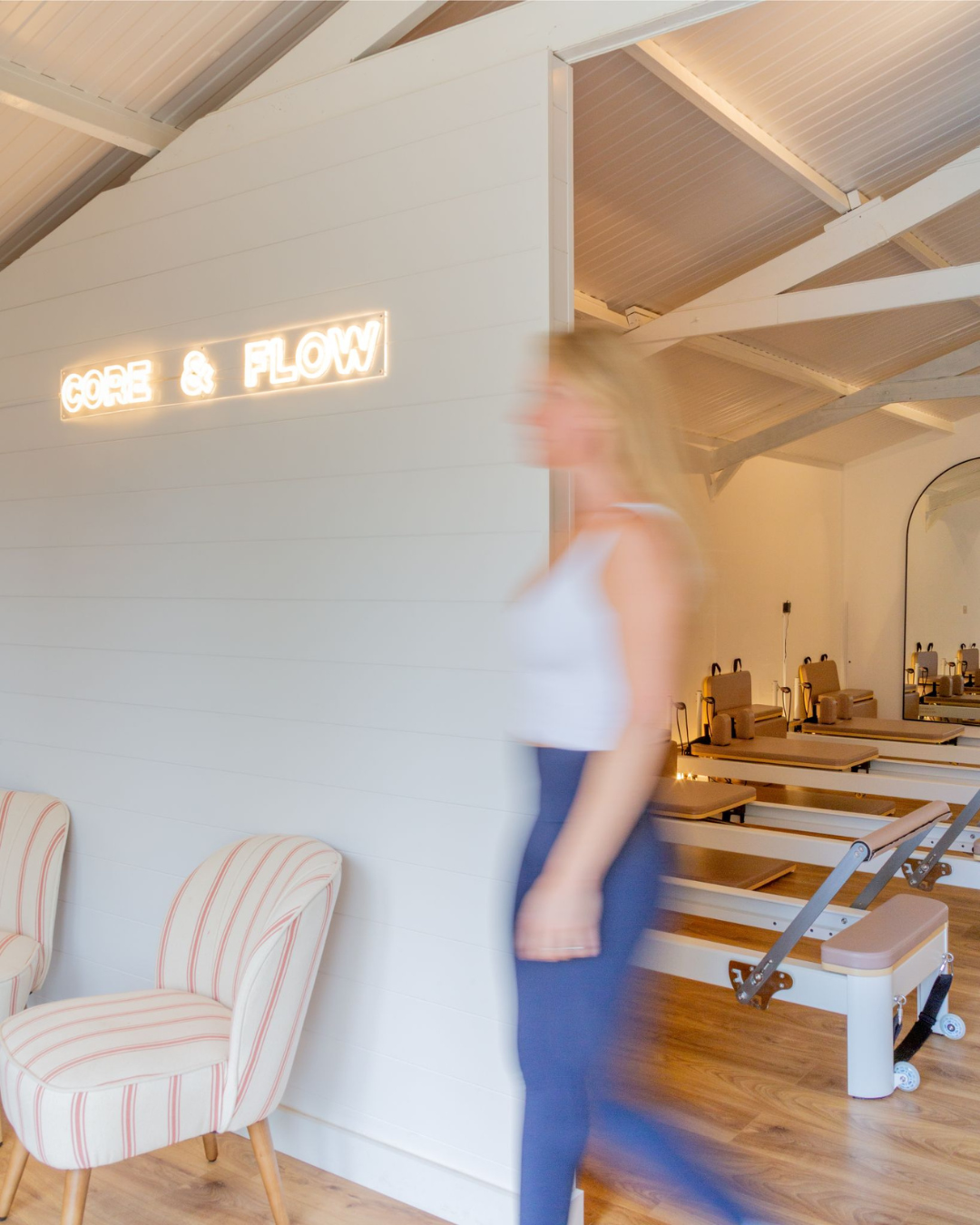Blurred woman walking past a wall with a neon sign that reads 'CORE & FLOW' in a wellness or fitness studio with pilates reformers and chairs.