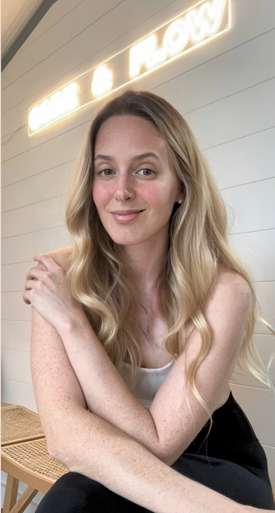 A woman with long blonde hair and freckles sitting indoors, smiling at the camera, with a glowing sign that says 'FLOW' in the background.