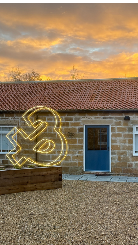 A yellow neon number 3 sign in front of a brick building with a blue door, set against a sunset sky.