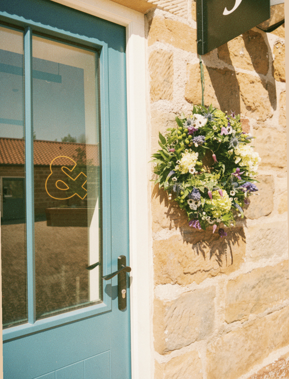 A blue door with a black handle and glass panel leading to Core & Flow Pilates studio, next to a stone wall with a hanging floral arrangement of white, purple, green, and black flowers.