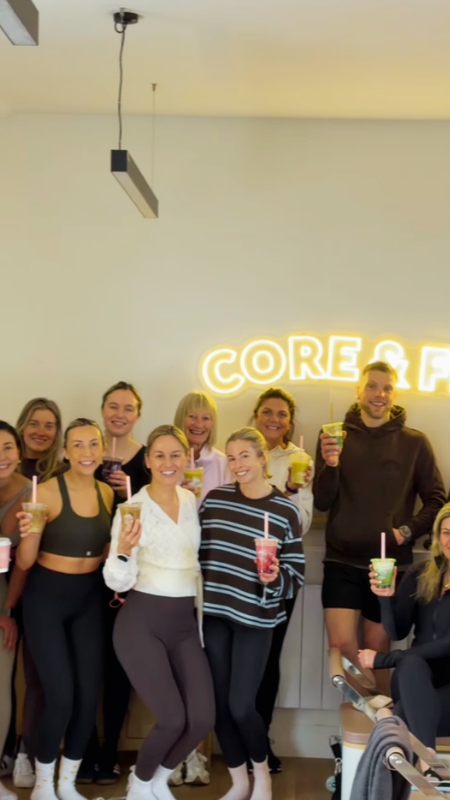 A group of people posing together in a Pilates studio, holding smoothies or drinks, in front of a wall with a neon sign that reads 'CORE & F'LOW.