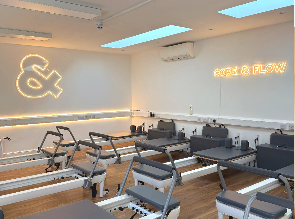 A Pilates studio with reformer machines arranged in rows on a wooden floor. The walls have neon signs, one with an ampersand symbol and another reading 'CORE & FLOW'. The room is well-lit with natural light from skylights and features white walls and a split air conditioning unit.