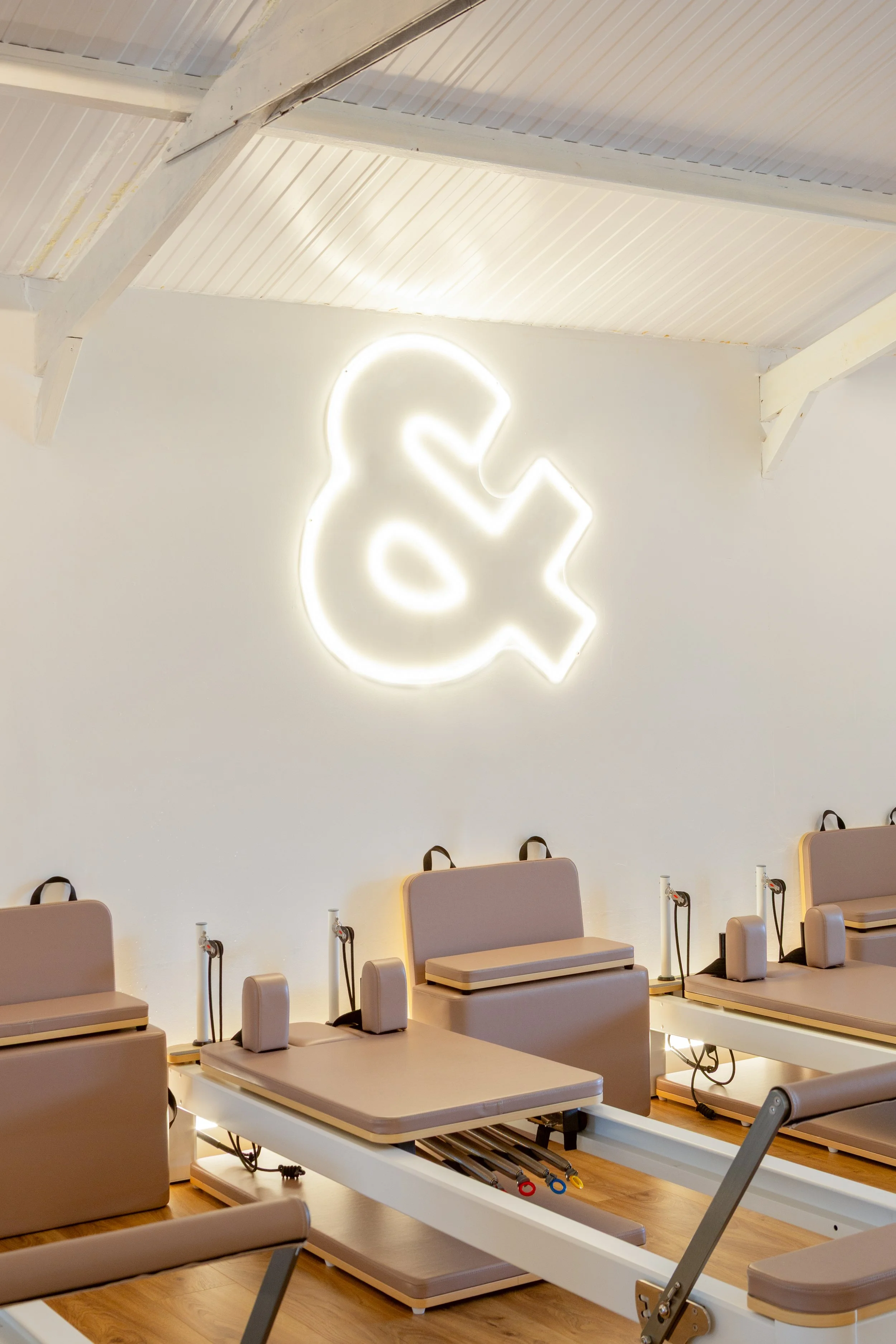 A Reformer Pilates studio full of Reformers and a white wall featuring a neon ampersand sign.
