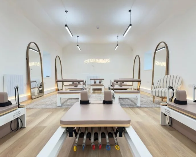 A modern Pilates or yoga studio with four reformer machines, large mirrors, and a cozy seating area with striped armchair, all illuminated by ceiling lights and ambient lighting.