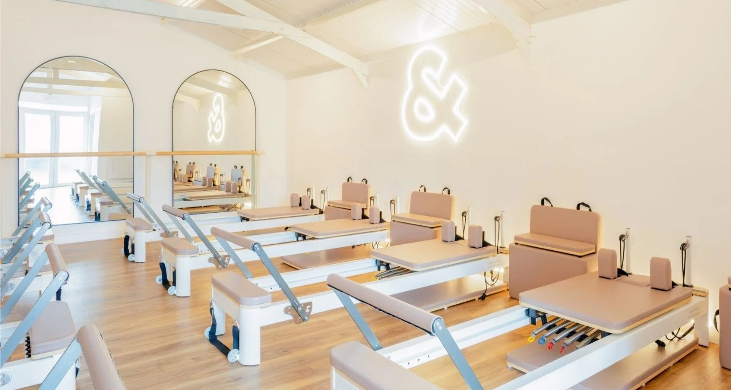 A Pilates or physical therapy studio with multiple reformer machines arranged in rows, large mirrors on the wall, and a neon sign of a ruffled fish on the wall.