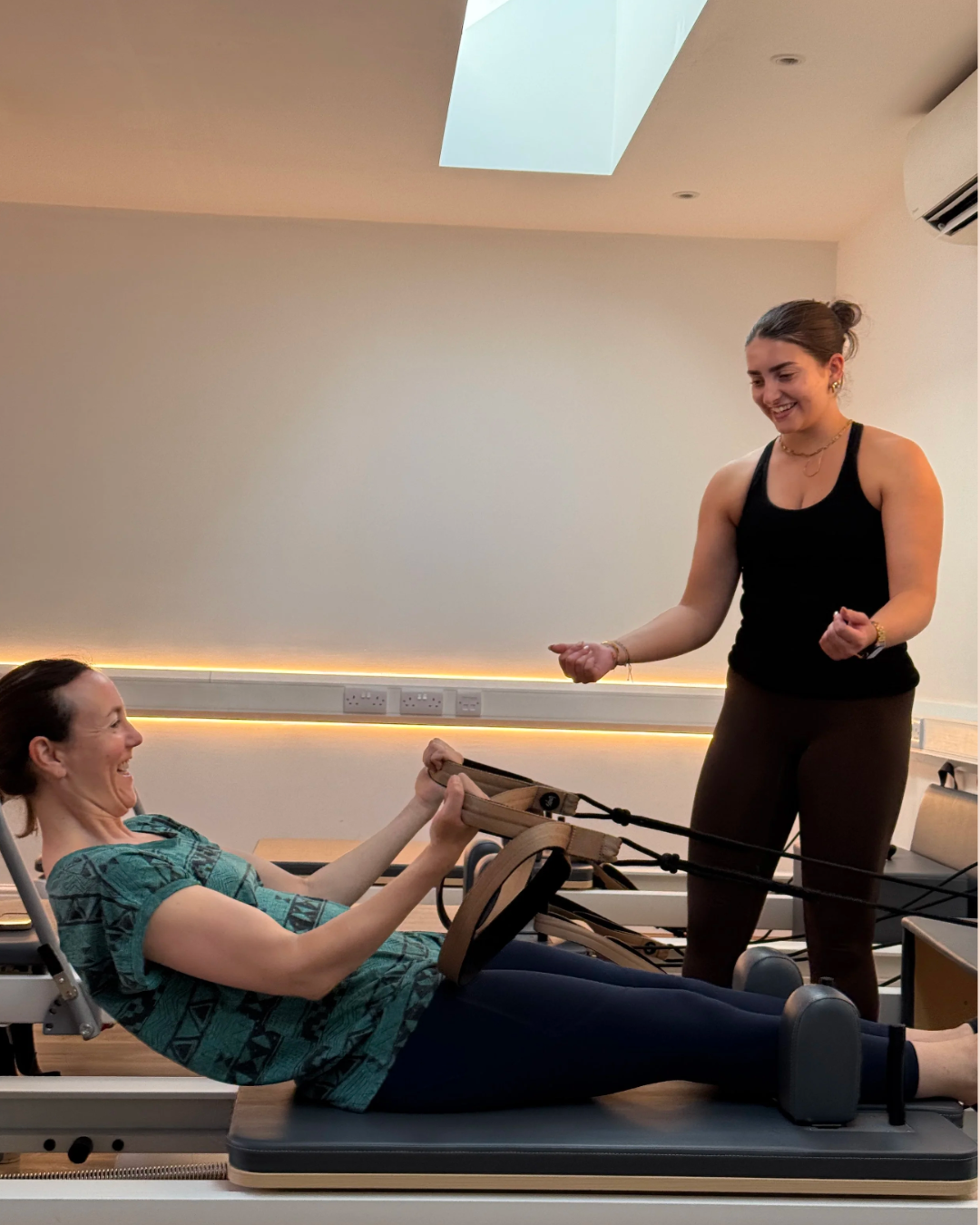 Two women in a Pilates Reformer studio, one lying on a Pilates reformer machine, smiling and holding the straps, the other standing beside, smiling and gesturing.