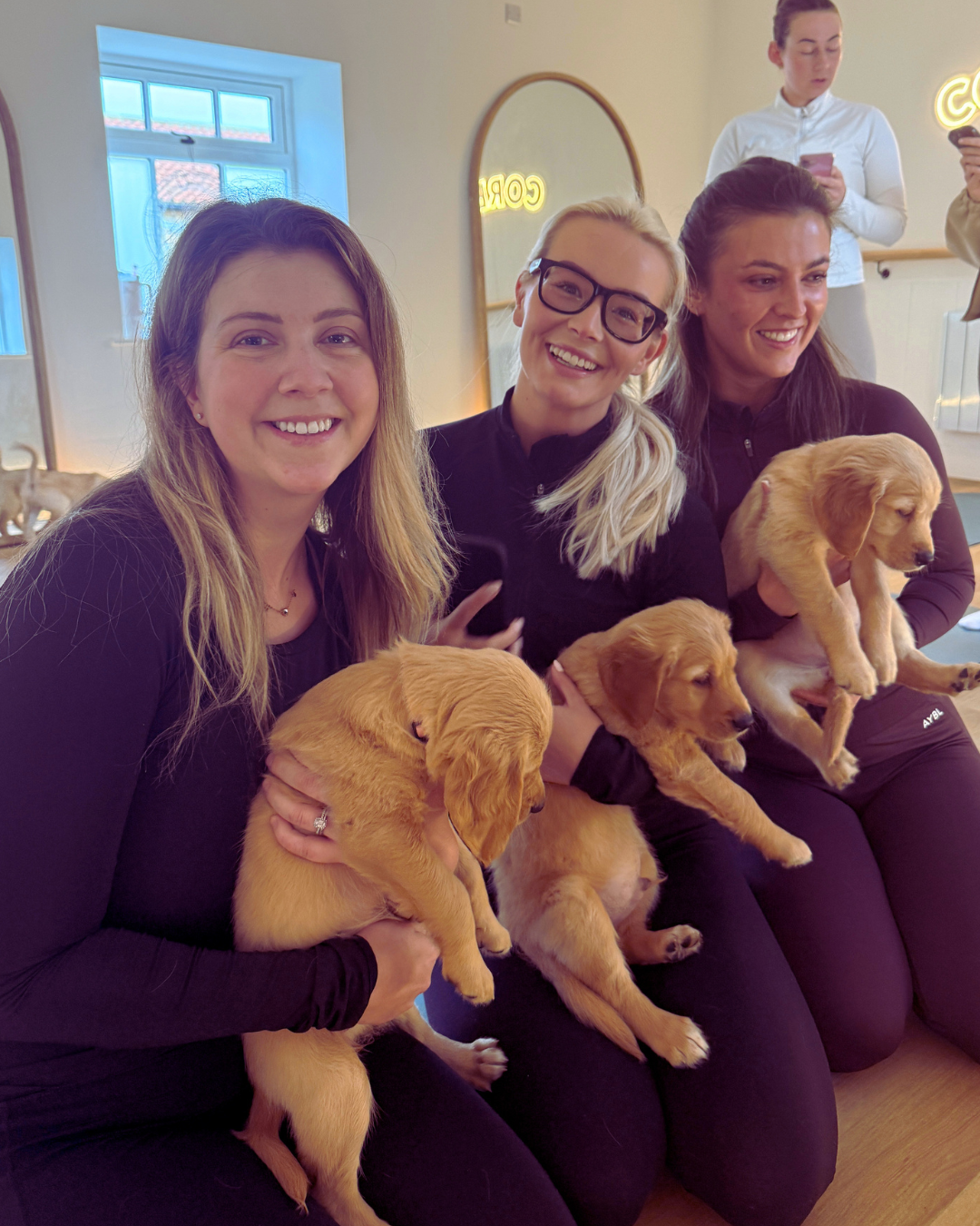 Three women sitting side by side smiling, each holding a yellow puppy in their arms, in a brightly lit indoor setting.