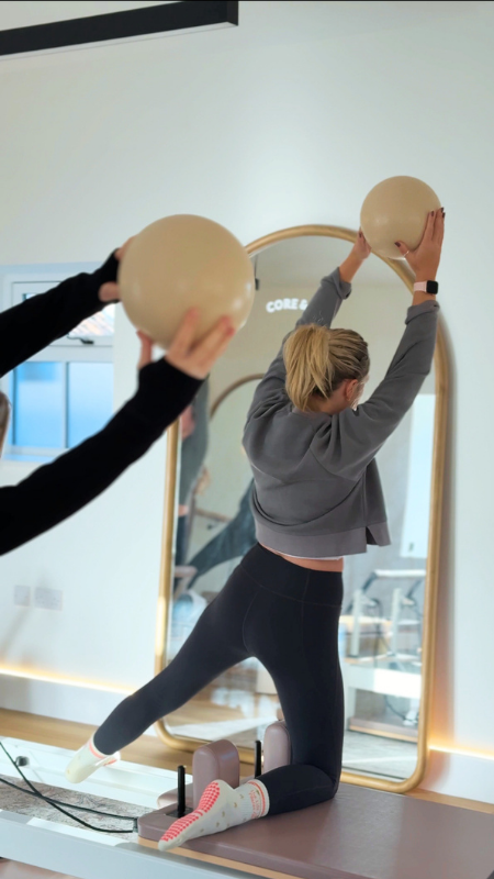Woman doing a wall stretch with a large exercise ball in a fitness studio, with her reflection visible in a mirror.