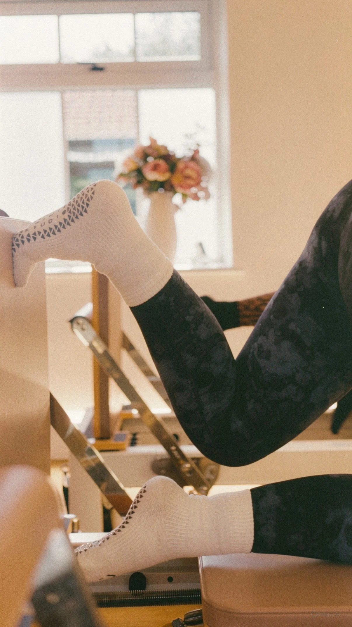 Person exercising on a Pilates reformer machine, wearing white socks and black athletic leggings, with a room containing large windows and a vase of flowers in the background.