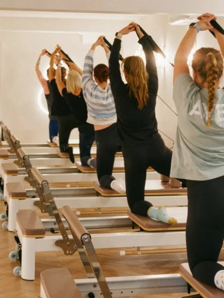 Group of women participating in a yoga or exercise class on reformer Pilates machines, facing away, in a studio with wooden floors and white walls.