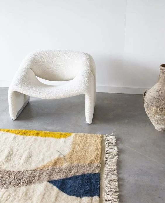 A white modern lounge chair, the Artifort M chair, with the iconic curved design, a large ceramic vase, and a colorful rug with fringe on a minimalist gray floor against a white wall.