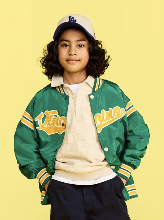 A bol brand image, showing a young boy with curly hair wearing a blue and white baseball cap, a green varsity jacket with yellow and white accents, a cream-colored polo shirt, and dark pants, standing against a yellow background.