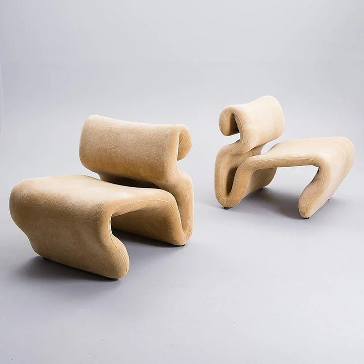 Two beige modern lounge chairs with abstract, curvy designs against a plain light gray background.