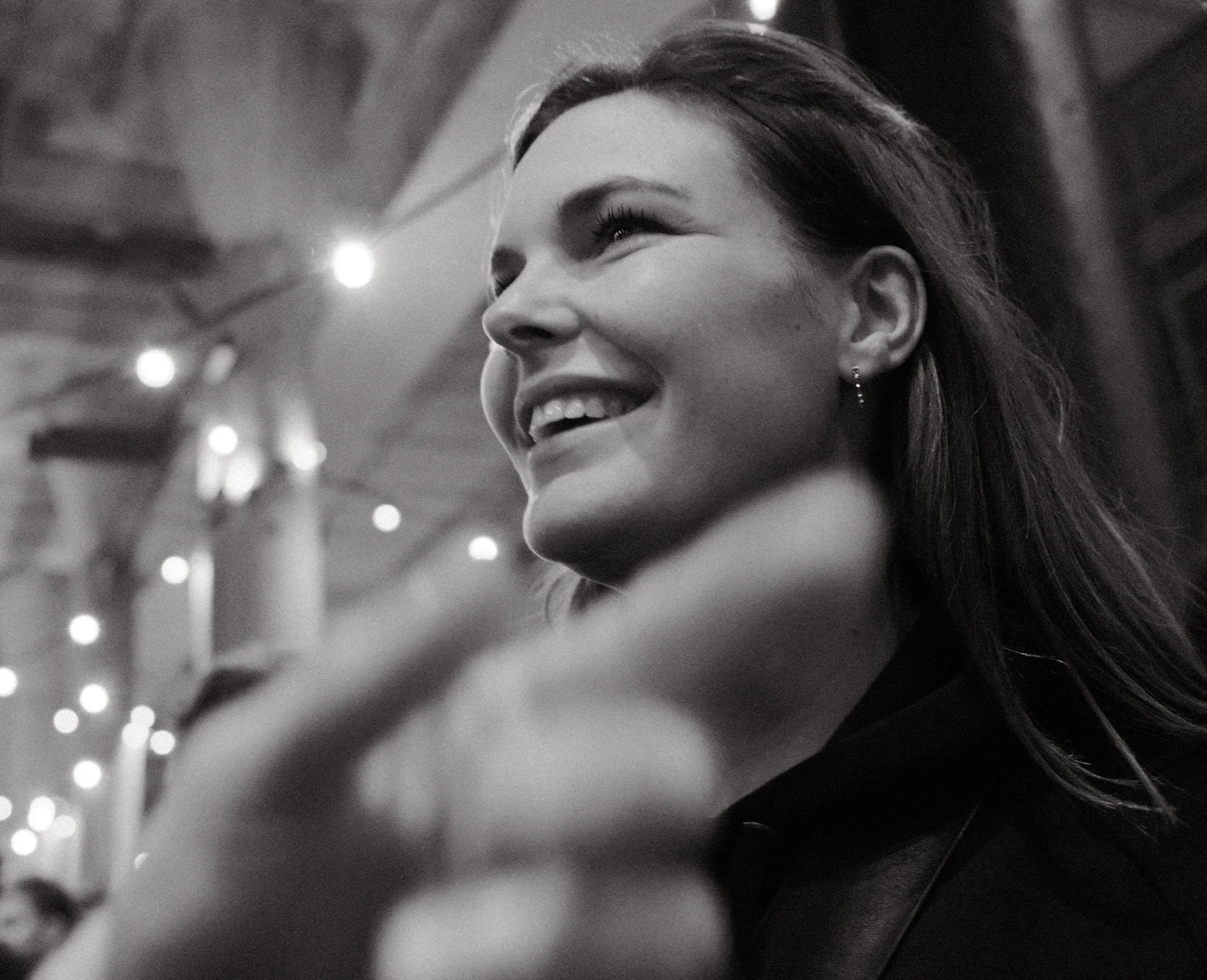 A smiling woman with long hair looking upward, with blurred lights in the background.