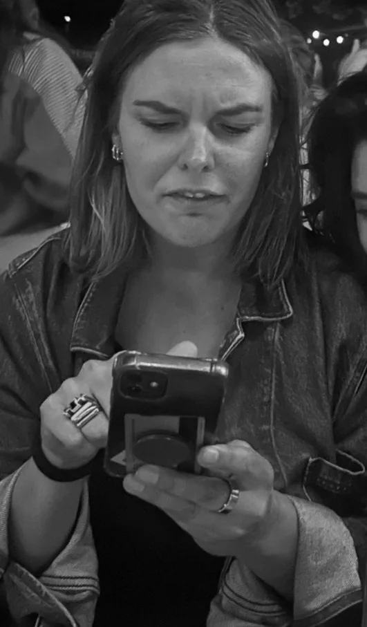A woman with shoulder-length hair, wearing a denim jacket, looking at her smartphone with a concerned or confused expression.