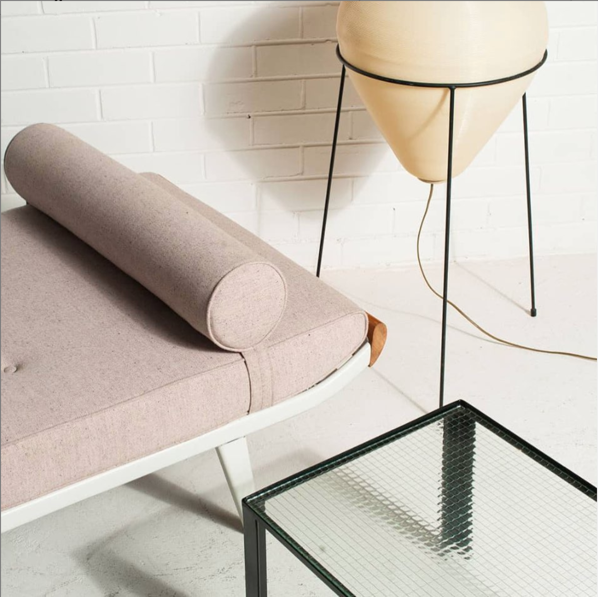 Interior of a modern living space with a pink upholstered Cleopatra daybed by Auping, a black metal side table with a glass top, and a tall, cream-colored planter on black wire legs against a white brick wall.