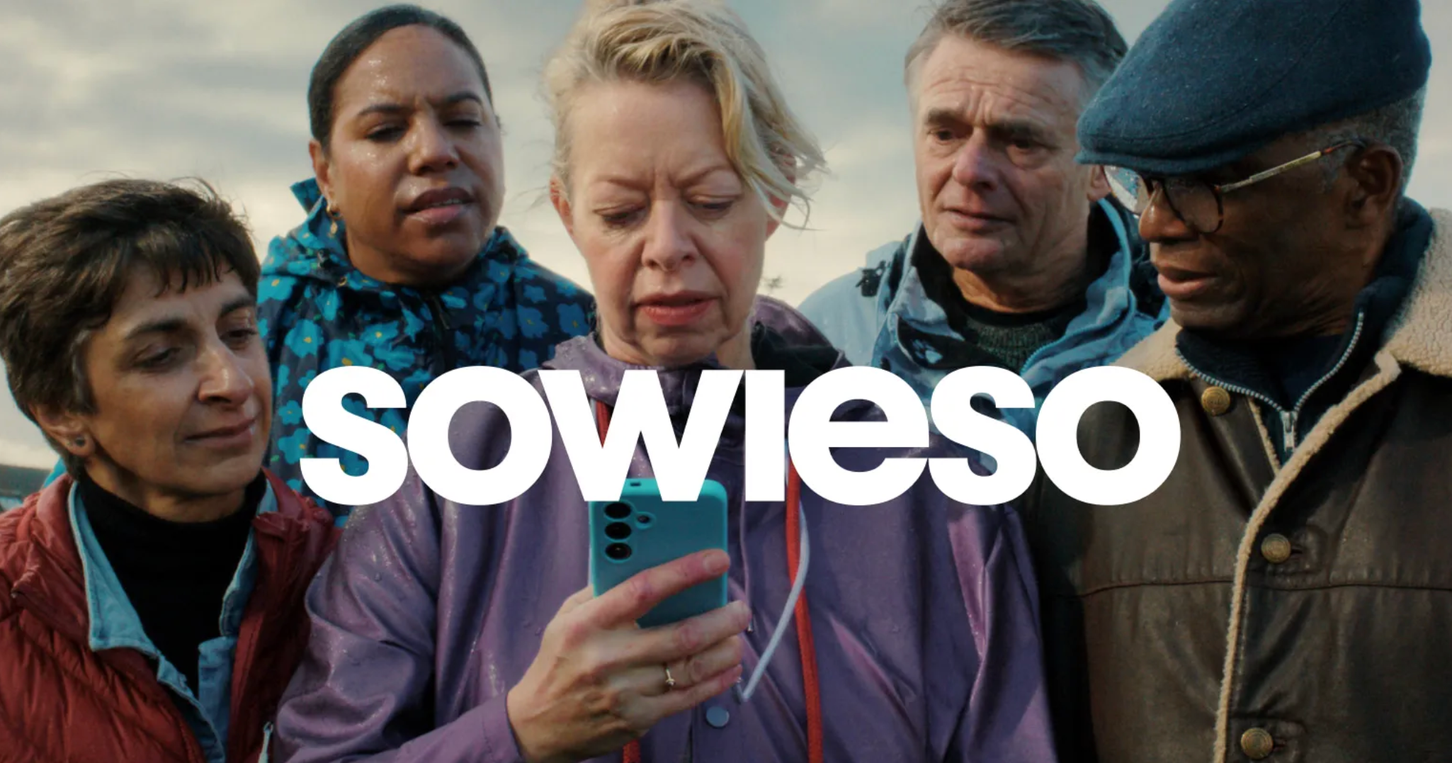 A group of six diverse people gathered closely outdoors, looking at a phone held by a woman in the center who is wearing a purple jacket. They appear to be focused or concerned, with cloudy sky in the background. The text 'SOWESO' is overlaid at the bottom of the image.