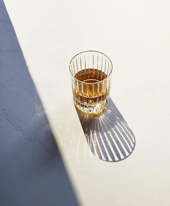 A glass of amber-colored liquid casting a shadow with a striped pattern on a white surface in sunlight.