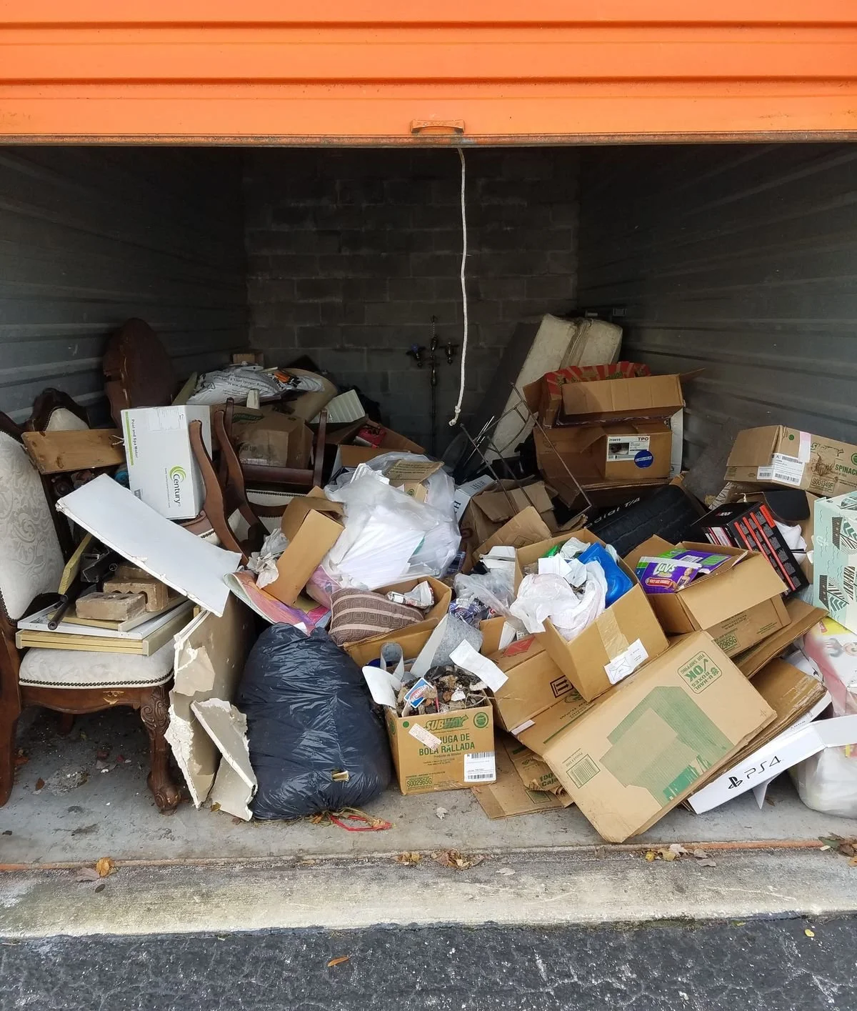 Overflow of discarded household items and trash in a garage with furniture, cardboard boxes, plastic bags, and debris.