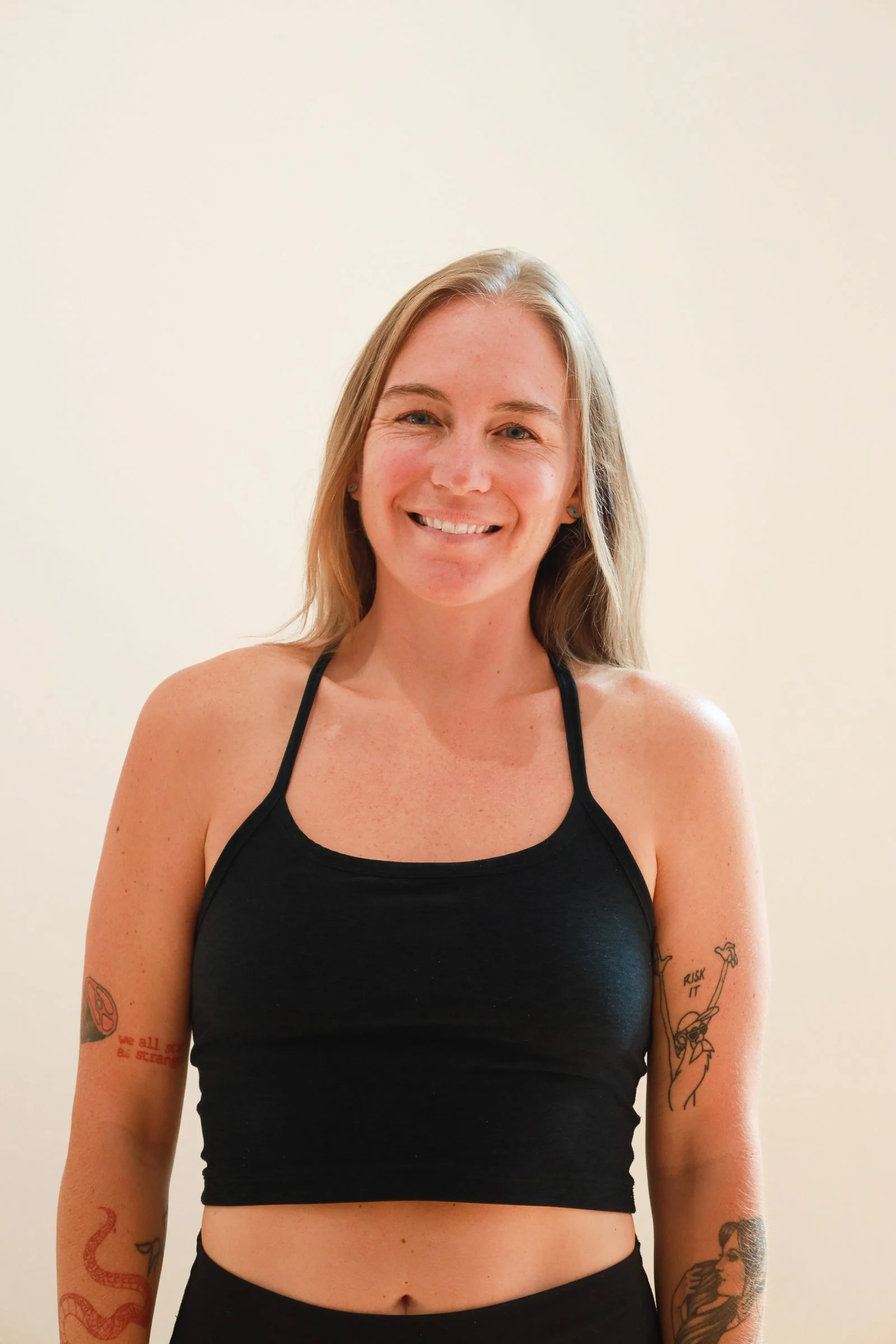 A smiling young woman with shoulder-length blonde hair, wearing a black crop top, standing against a plain, light-colored background.