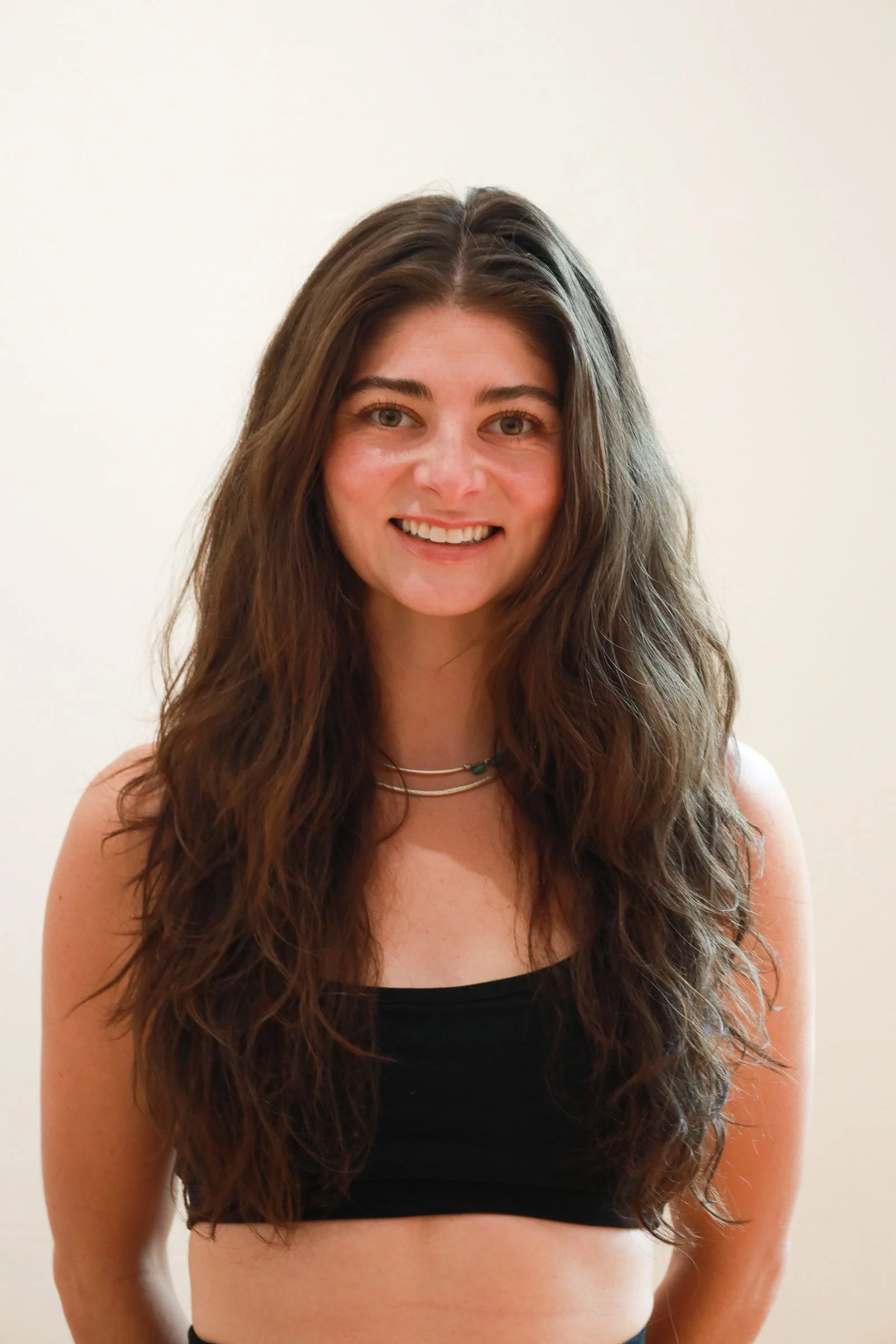 A young woman with long, wavy brown hair, smiling, wearing a black tube top and layered necklaces, standing against a plain white wall.
