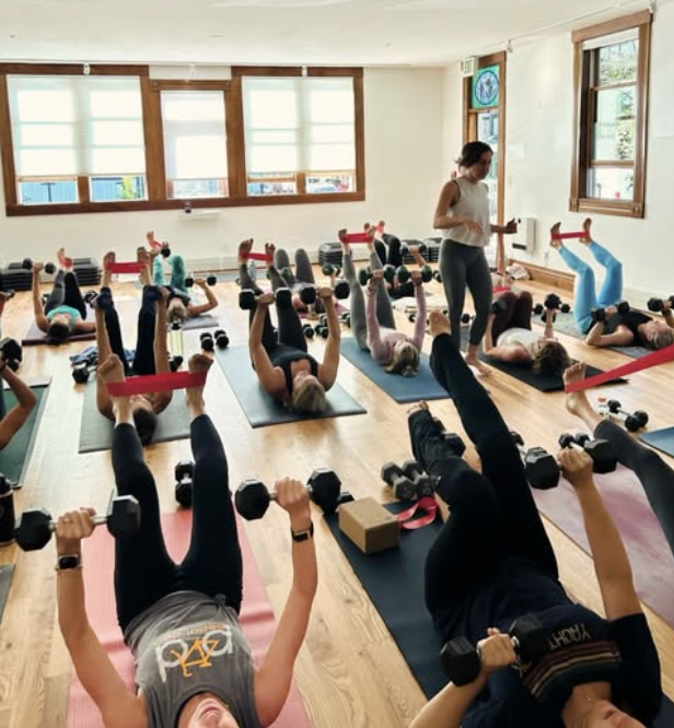 A group fitness class at Range Yoga with participants lying on yoga mats, lifting dumbbells and holding resistance bands, led by a woman instructor in a bright, wooden-floored room with large windows.