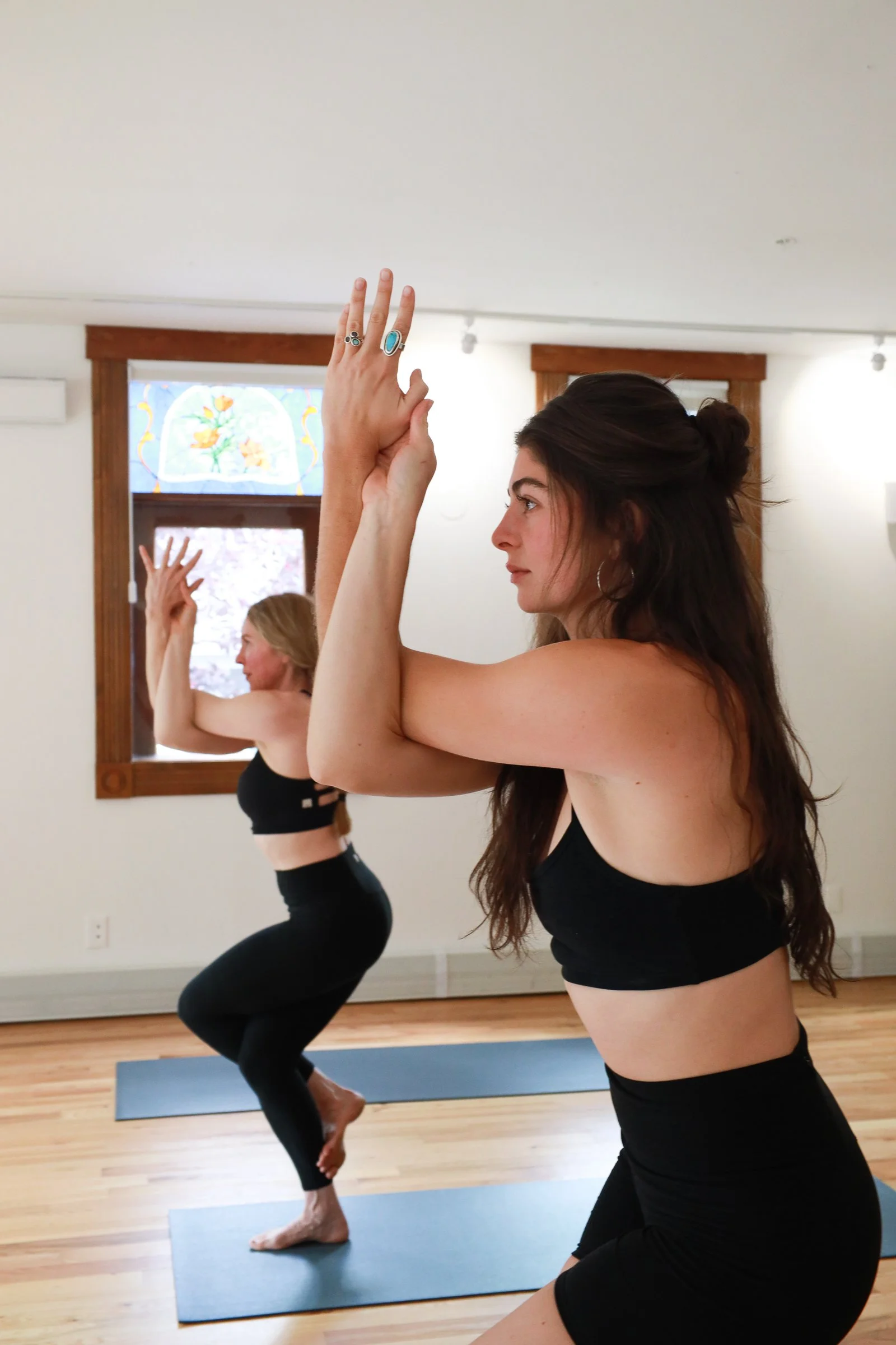 Two women practicing yoga indoors at the Range Yoga Studio in Telluride, Colorado. They are in eagle pose. The woman in the foreground has long brown hair in a bun, and the woman in the background has blonde hair.