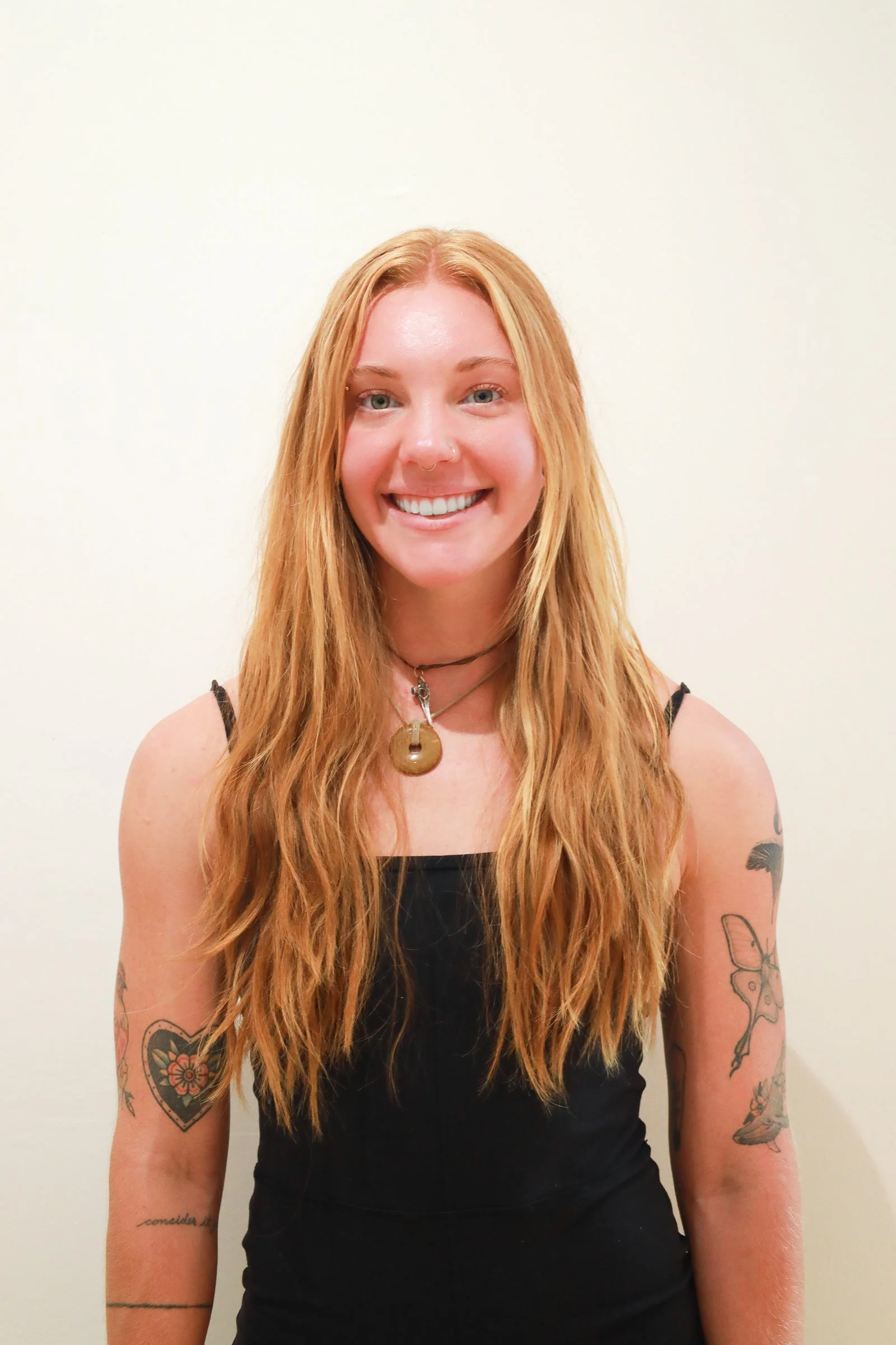 A young woman with long, wavy red hair and blue eyes, smiling, wearing a black spaghetti strap top, and tattoos on her left arm and right forearm, standing against a plain white background.